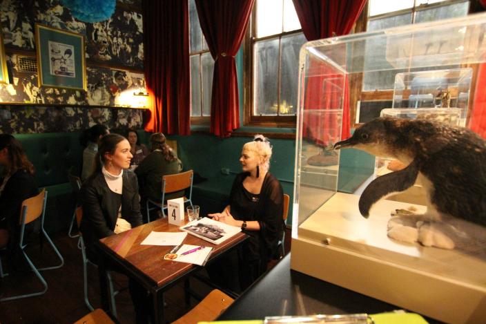 "Two young women talking at a table, taxidermied penguin in a perspex case in the foreground."